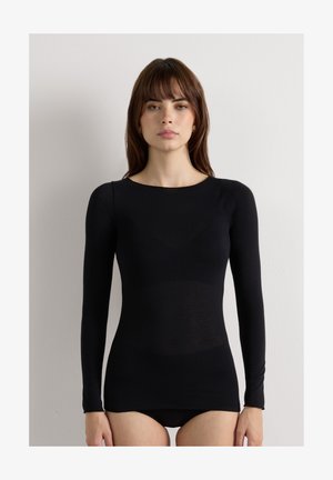 Femme aux cheveux bruns lisses portant un haut noir moulant à manches longues, debout contre un mur clair et uni, faisant face.