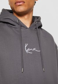 Sudadera gris con cordón y puntas de metal. Presenta texto bordado en blanco en el pecho. La tela parece suave y tiene una textura lisa.