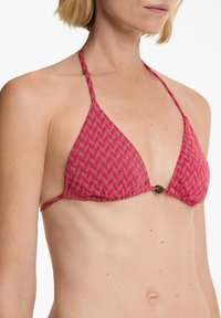 Rdeča in bela vzorčasta trikotna bikini zgornji del s tankimi naramnicami in osrednjo kovinsko okrasitvijo, ki prikazuje teksturirano tkanino.