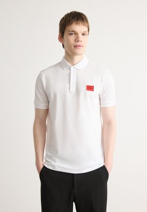 Weißes Poloshirt mit kurzen Ärmeln, das einen Kragen, eine Dreiknopfleiste und ein rotes Markenpatch auf der Brust umfasst. Glatte Baumwolltextur.