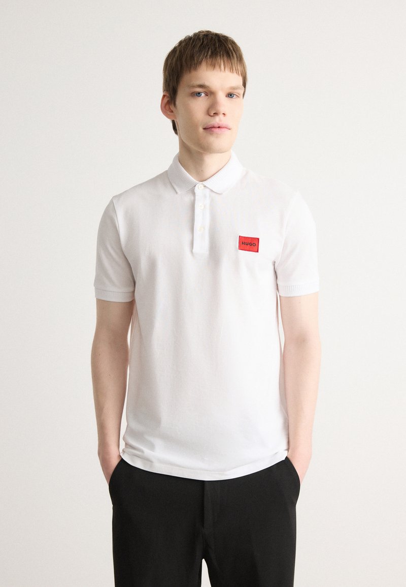 Hvid kortærmet polo-shirt med krave, tre knaplukninger og et rødt mærke på brystet. Glat bomuldsstruktur.