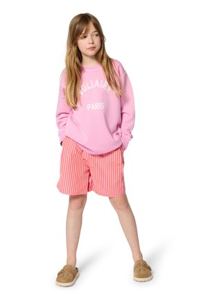 Fille debout, les mains dans les poches, portant un sweat-shirt rose avec le texte "PARIS", un short rayé orange et des chaussures marron sans lacets.