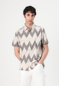 Camicetta a maniche corte in tessuto beige con un motivo a zigzag grigio scuro. Presenta un colletto e una chiusura a bottoni, abbinata a pantaloni bianchi.