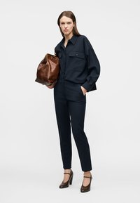 Femeie îmbrăcată într-o cămașă neagră cu nasturi și pantaloni, ținând o geantă mare de piele maro, stând cu o mână în buzunar, pe fundal alb.