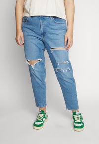 Personne portant un jean bleu clair déchiré et des baskets vertes et blanches, debout sur un fond clair uni.