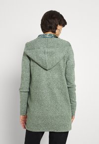 Dlouhý zelený kardigan s kapucí z texturované pletené látky. Má kontrastní límec a prodloužený zadní lem.