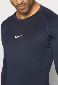 Marineblaues Sportoberteil mit langen Ärmeln aus elastischem Material, mit einem weißen Nike-Logo auf der Brust und kontrastierenden Nähten.