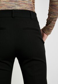 Gros plan d'une personne portant un pantalon noir sur mesure et un haut texturé à manches longues, une main dans une poche sur un fond uni.