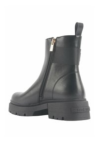 Bota negra de cuero hasta el tobillo con cremallera lateral, detalle de banda elástica y suela gruesa con relieve. Presenta un acento de logo en el talón.