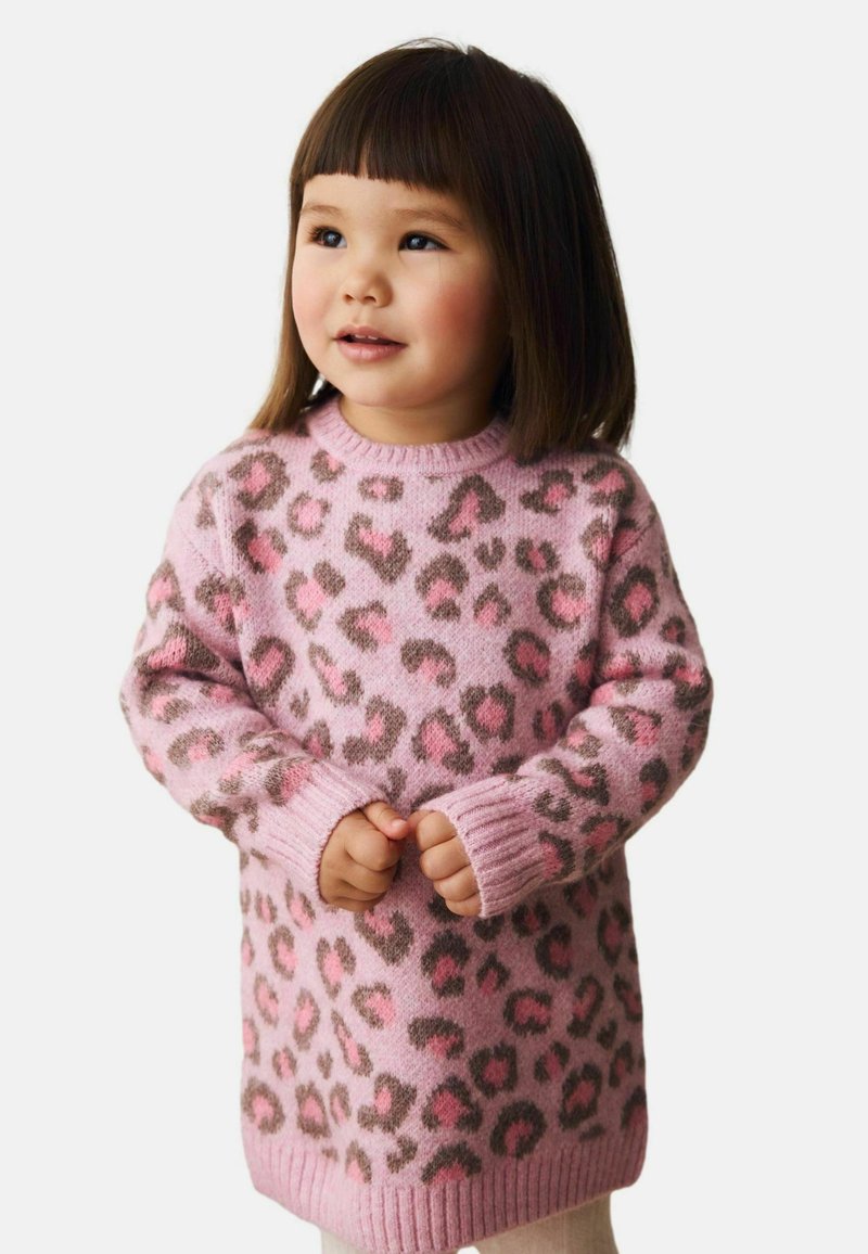 Junges Mädchen mit glattem braunem Haar, das einen rosa Leopardenmuster-Pullover trägt, steht und schaut leicht zur Seite.