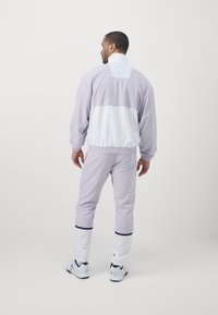 Lilafarbener Trainingsanzug mit einem weißen Einsatz im Rücken und an den Bündchen; verfügt über einen elastischen Bund und Bündchen, mit kontrastierenden marineblauen Akzenten und weißen Sneakers.