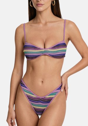 Donna che indossa un bikini a righe viola, blu, verde e giallo con spalline sottili, posando con una mano sul fianco davanti a uno sfondo semplice.