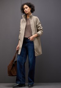 Beżowy płaszcz z wysokim kołnierzem, strukturalny brązowy sweter, szerokie ciemnoniebieskie dżinsy i czarne botki za kostkę, trzymając brązową torebkę z zamszu.