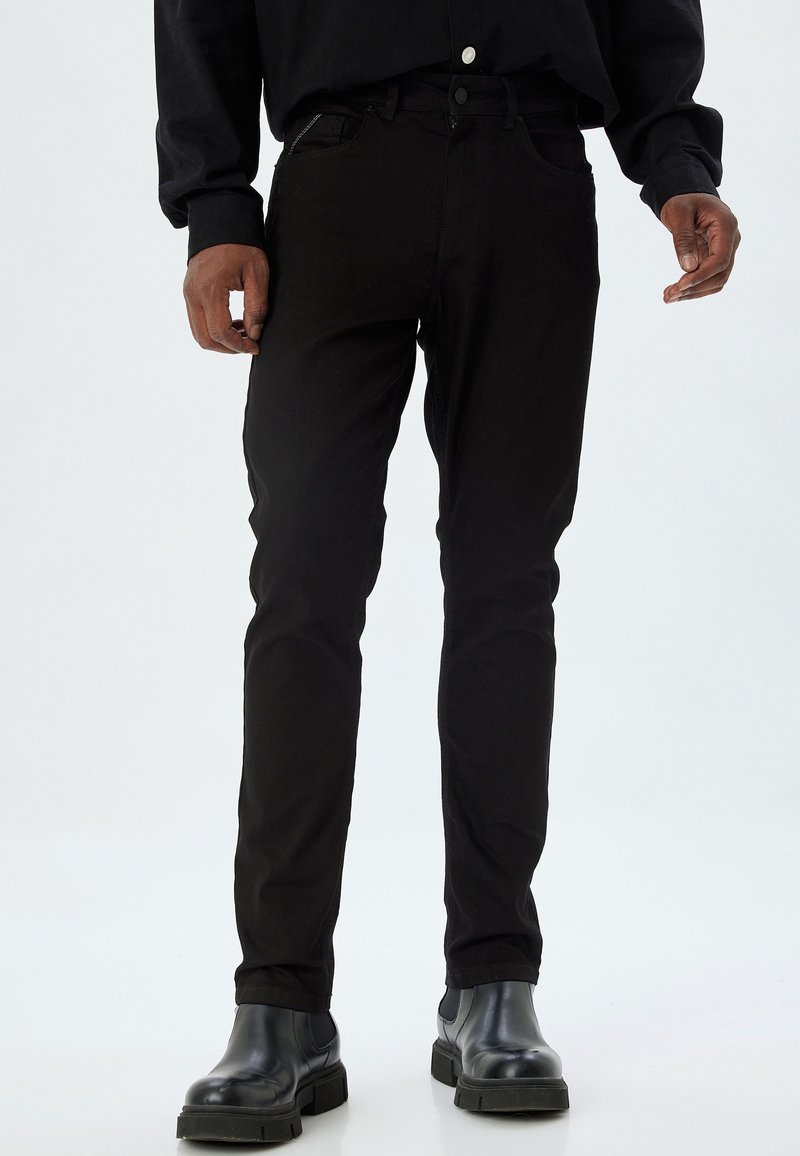 Mustat farkut slim fit -mallina, joissa on klassinen viisi taskua -muotoilu, sävyyn ommeltu saumaus ja nappikiinnitys. Yhdistetty mustiin saappaisiin.