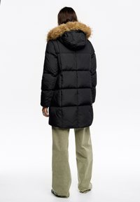 Czarna pikowana kurtka puchowa z kapturem wykończonym futrem, o przyszytym wzorze i nieco dłuższym kroju, zestawiona z jasnozielonymi szerokimi spodniami.