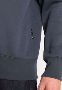 Grå sweatshirt med ribbade ärmslut och nederkant, med en G-Star-tag på sidan. Slät textur, avslappnad passform och rektangulär design.