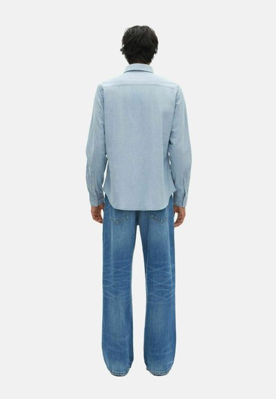 Camicia azzurra chiaro con colletto e orlo curvo, abbinata a jeans blu a gamba larga con tasche posteriori e una texture sbiadita.