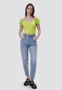 Bodysuit verde lima con mangas con volantes, combinado con unos jeans de tiro alto en azul claro. El modelo lleva sandalias negras gruesas.
