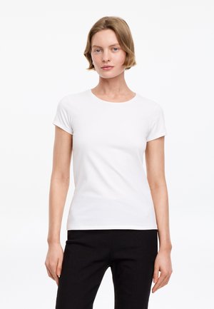Femme aux cheveux courts châtain clair portant un t-shirt blanc uni à manches courtes et un pantalon noir, debout devant un fond blanc.