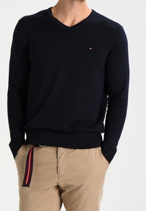 Mann trägt einen schwarzen V-Ausschnitt-Pullover und beige Hose, die Hände in den Taschen, und ein gestreifter Stoffgürtel hängt von der Taille.