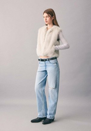 Veste en fausse fourrure blanche courte, haut à manches longues gris clair, jean en denim bleu clair déchiré avec une ceinture noire cloutée, et bottines noires. 