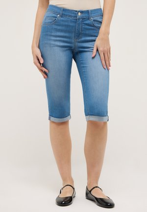Kvinde iført midtblå rullede denim bermuda-shorts og sorte Mary Jane-flade sko, stående mod en ensfarvet baggrund.