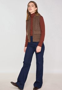 Gilet di maglia marrone con due tasche frontali sopra un dolcevita aderente bordeaux; abbinato a pantaloni a campana blu navy e scarpe marroni.