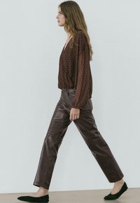 Frau mit langen Haaren geht, trägt eine braune Bluse mit weißen Punkten, dunkelbraune Lederhose und schwarze Ballerinas vor einem einfarbigen Hintergrund.