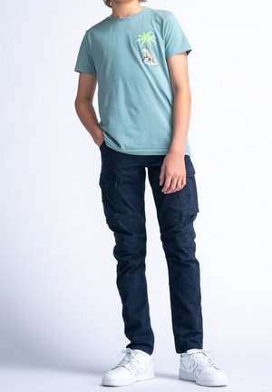 T-shirt in cotone azzurro chiaro con grafica di una palma sul petto, abbinata a pantaloni cargo blu navy e sneakers bianche. Abbigliamento casual per giovane.