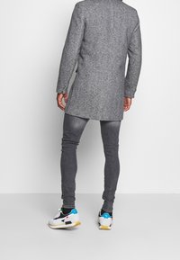 Grå uldblandet frakke med en lang silhuet, parret med mørkegrå tætsiddende jeans og hvide sneakers med sorte og teal accenter.