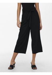 Pantalons larges noirs à taille haute, dotés d'une ceinture en tissu. Texture lisse, design plissé et longueur cropped. Portés avec des mules à talons noirs.