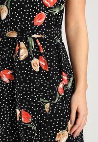 Černé šaty s bílými puntíky, zdobené červenými a béžovými květinovými vzory. Obsahují šňůrku v pase a hladkou texturu látky.