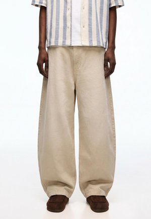 Persoon die beige wijde broek draagt, bruine schoenen en een blauw-wit verticaal gestreept overhemd met knopen over een wit hemdje.