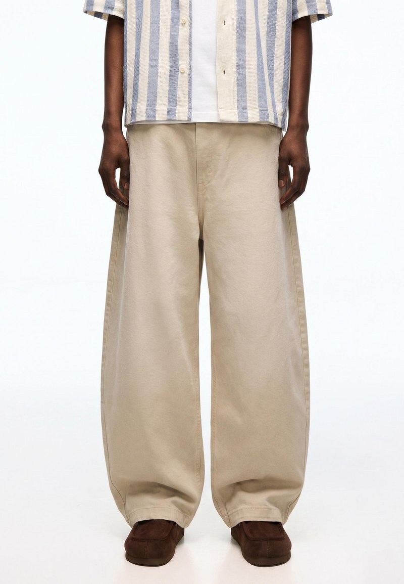 Personne portant un pantalon beige à jambes larges, des chaussures marron et une chemise à rayures verticales bleu et blanc boutonnée sur un haut blanc.