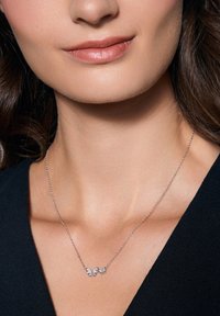 Srebrny naszyjnik w kształcie motyla z trzema zdobionymi zawieszkami, ozdobiony małymi przezroczystymi kamieniami szlachetnymi i delikatnym łańcuszkiem. Noszony na ciemnym tle.