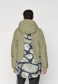 Olive green jacket with a patterned navy and light blue back, featuring an adjustable hood and multiple pockets. Made from textured material.