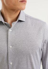 Man som bär en ljusgrå, strukturerad skjorta med knappar, med kragen öppen, visad från hakspets till bröstet mot en enfärgad bakgrund.