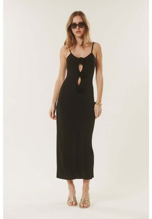 Femme portant une robe midi noire sans manches avec des découpes sur le devant et des liens floraux, des sandales dorées, des lunettes de soleil et un bracelet doré, debout sur fond blanc.