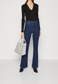 Haut noir côtelé à manches longues avec un col en V, associé à un jean denim bleu foncé évasé. Le mannequin tient un sac fourre-tout blanc à rayures et porte des bottes noires.