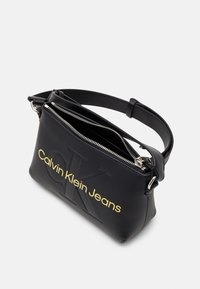 Černá kabelka z umělé kůže s texturovaným povrchem, zdobená zlatým potiskem loga, se dvěma zipovými přihrádky a nastavitelným ramenním popruhem.