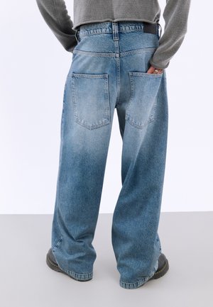 Personne portant un jean bleu ample et un pull gris, avec une main dans la poche arrière, debout sur un sol gris.