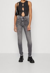 Jeans de tiro alto en gris combinados con un bodysuit negro sin mangas que presenta detalles recortados y una textura suave. Usados con botas negras gruesas.
