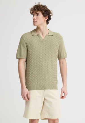 Młody mężczyzna o kręconych włosach, ubrany w teksturowaną oliwkowozieloną krótko rękawową dzianinową koszulkę polo i beżowe szorty do kolan, stojący na jednolitym tle.