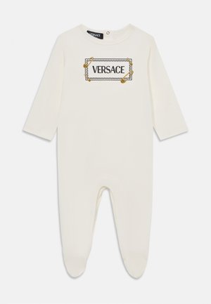 Krémovo sfarbený detský overal vyrobený z jemného materiálu, s dlhými rukávmi, zaobleným výstrihom a čierno-zlatou potlačou loga "VERSACE".