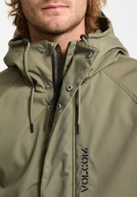 La veste à capuche verte possède une fermeture éclair à l'avant, des boutons-pression, des cordons ajustables et un logo noir sur le côté. Texture de tissu douce et mate.