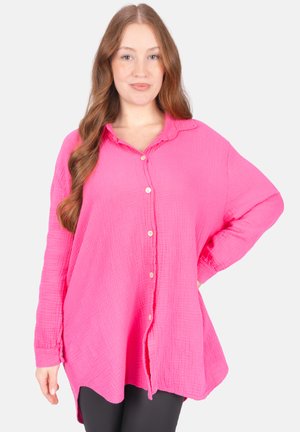 Woman with long wavy hair wearing a bright pink button-up textured blouse and black pants, standing with one hand on hip against white background.