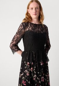 Juodas nėrinių suknelė su permatomomis ilgomis rankovėmis, gėlių raštuotu sijonu, peplum juosmens detalėmis ir sudėtingais nėriniais, demonstruojanti tekstūrų derinį.