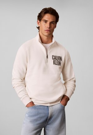 Junger Mann trägt einen cremefarbenen Calvin Klein Viertel-Zip-Sweatshirt und hellblaue Jeans, steht mit den Händen in den Taschen vor einem einfarbigen Hintergrund.