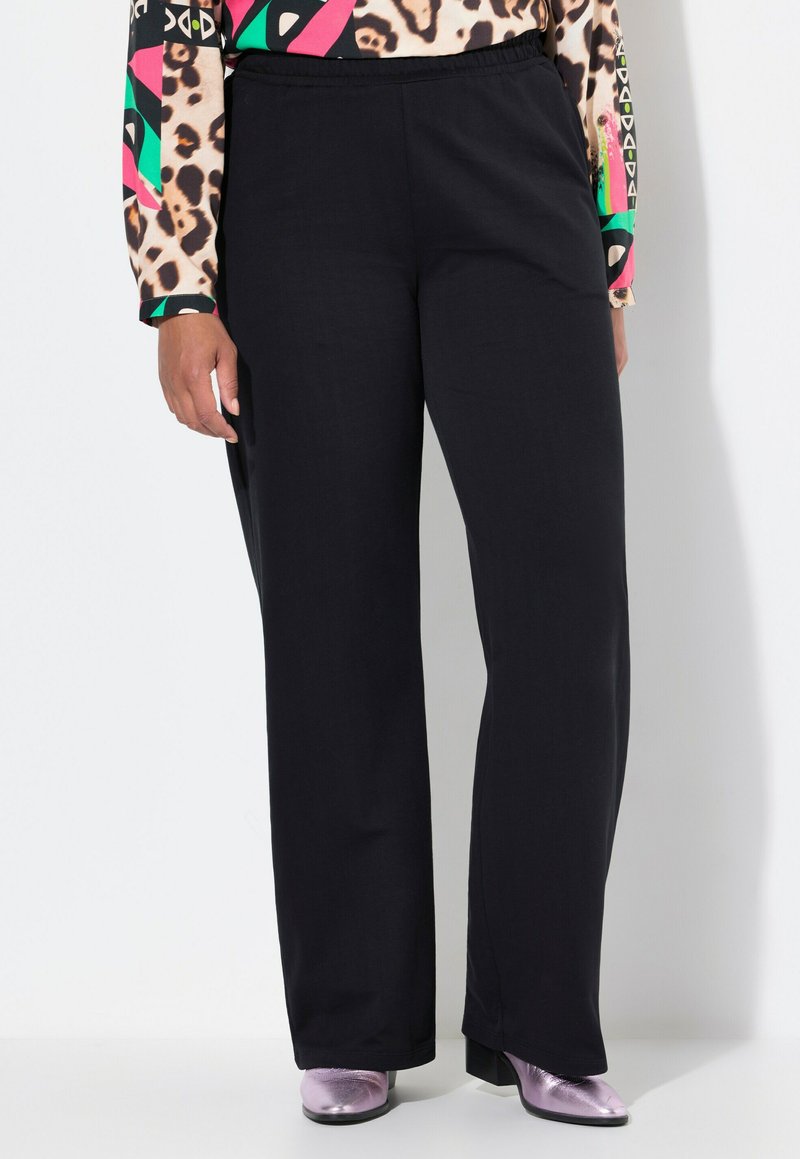 Zwarte, brede broek met een elastische tailleband en een gladde textuur, gecombineerd met glanzende roze enkellaarsjes. De achtergrond is een lichte neutrale tint.