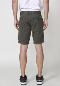 Vīrietis, kurš valkā sarullētas tumši zaļas šortus, baltu t-kreklu un melnus sporta apavus, stāv ar muguru pret vienkāršu gaišu fona.